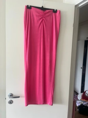 Lång rosa kjol med rynkdetalj - En lång, rak kjol i stark rosa färg med snygg rynkdetalj framtill i midjan. Kjolen har en enkel och stilren design som passar perfekt till sommaren. Materialet känns mjukt och följsamt.