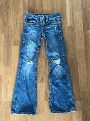 Blå bootcut jeans med slitningar - Säljer ett par blå bootcut jeans med slitna detaljer och hål på knäna. Jeansen har klassisk femficksdesign, knappgylf och kontrastsömmar. Materialet är denim i bomull. Perfekta för en avslappnad och trendig look.