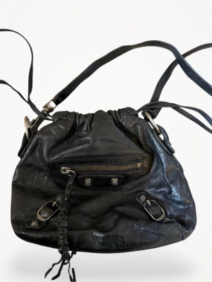 Svart axelremsväska från Balenciaga - Cool svart handväska från Balenciaga i glansigt läder med justerbar axelrem, dragkedjeficka framtill och dekorativa metalldetaljer. Väskan har en avslappnad bucket-form och flätad dragkedjedekor som ger en edgy vibe.  Äkta!!