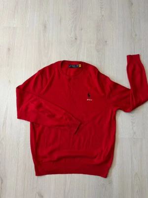 Röd tröja från Polo Ralph Lauren - Riktigt snygg röd tröja från Polo Ralph Lauren med klassisk rund hals och broderad logga på bröstet. Tröjan är långärmad och har ribbade muddar vid ärmslut och nederkant. Perfekt för dig som gillar stilrena och tidlösa plagg.