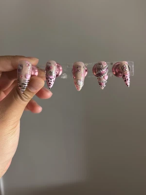 Rosa Hello Kitty press-on naglar - Supergulliga press-on naglar i rosa med Hello Kitty-motiv, svarta ränder, prickar och små hjärtan. Långa och spetsiga med glittriga detaljer och 3D-effekter. Perfekt för dig som vill ha en söt och unik look. Material: akryl.