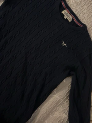Mörkblå kabelstickad tröja - Mörkblå kabelstickad tröja  med rund halsringning och broderad logga på bröstet. Tröjan har ribbade muddar och klassisk passform. Tillverkad i mjukt material som passar perfekt till lager på lager.
