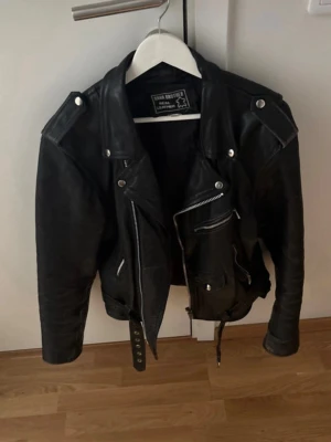 Svart biker skinnjacka XL - Cool svart biker skinnjacka med klassiska silvriga dragkedjor, axelklaffar och bälte i midjan. Jackan har flera fickor med dragkedja och en snygg asymmetrisk front. Perfekt för en edgy streetstyle-look.