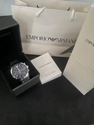 Emporio Armani silverfärgad klocka blå - Stilren klocka från Emporio Armani med silverfärgat armband i metall och mörkblå urtavla. Klockan har kronograf-funktion, tydliga index och märkets logga vid tolvslaget. Perfekt accessoar för dig som gillar klassisk och modern design.