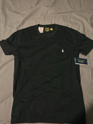 Svart t-shirt från Polo Ralph Lauren - Svart t-shirt från Polo Ralph Lauren med klassisk passform och liten vit logga broderad på bröstet. T-shirten har rund halsringning och är kortärmad, perfekt för en stilren och enkel look.