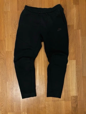 Nike tech byxor - Svarta mjukisbyxor från Nike med dragsko i midjan och ribbade muddar vid bensluten. Byxorna har en slim fit-modell och är tillverkade i mjukt material med Nike-logga tryckt på låret. Perfekta för en avslappnad och sportig stil. De är barn storlek XL men passar även XS och S i herr (upp till 175cm) 