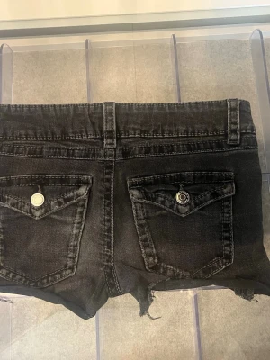Svarta jeansshorts med råa kanter - Svarta denimshorts med råa, fransiga kanter och klassisk femficksdesign. Shortsen har bälteshällor, knapp- och dragkedjestängning samt slitna detaljer för en cool look. 