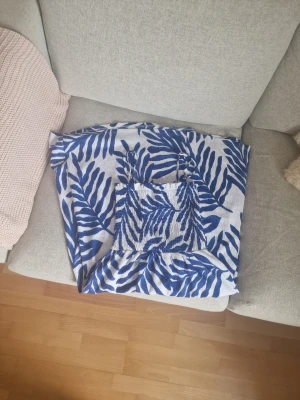 Blåvit sommarklänning med mönster - Säljer en blåvit klänning med breda axelband och tropiskt mönster. Klänningen har smockad överdel och volang nertill, perfekt för varma dagar. Lätt och luftig modell som ger en avslappnad känsla.