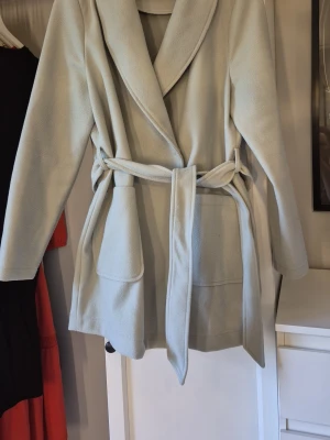 Ljusgrön kappa med knytskärp - Stilren ljusgrön kappa med bred krage och knytskärp i midjan. Kappan har två stora fickor framtill och är tillverkad i ett mjukt tyg som ger en avslappnad men ändå elegant look. Är lite nopprig på vissa ställen, är av sånt material så det blir lätt så. Katt finns I hemmet.