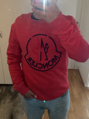 Röd Moncler sweatshirt med logga - Röd sweatshirt från Moncler med stor broderad logga i mörkblått på bröstet. Tröjan har rund hals, långa ärmar och ribbade muddar vid ärmslut och nederkant. Perfekt för en trendig och avslappnad stil.