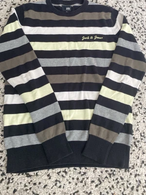 Randig tröja från Jack & Jones - Snygg långärmad tröja från Jack & Jones med breda horisontella ränder i svart, grått, vitt, beige och ljusgul. Tröjan har rund halsringning och broderad logga på bröstet. Perfekt för en avslappnad stil.