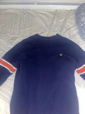Blå sweatshirt från Lyle & Scott - Mörkblå sweatshirt från Lyle & Scott med rund hals och klassisk gul logga på bröstet. Tröjan har vita och röda ränder på ärmarna för en sportig look. Perfekt för dig som gillar stilrena och bekväma plagg. Nypris 600kr