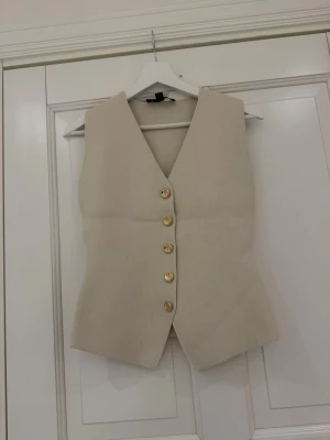 Beige väst med guldknappar - Stilren beige väst i ribbat tyg med två guldknappar framtill. Västen har en figurnära passform och är ärmlös med klassisk krage. Perfekt att styla över en skjorta eller t-shirt för en trendig look.