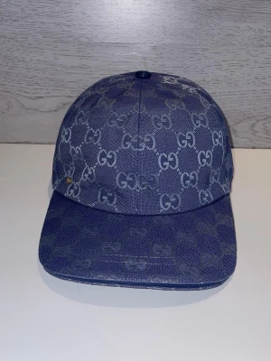 Blå denimkeps  - Snygg blå keps i denim med det klassiska mönstret över hela kepsen. Böjd skärm och justerbar baksida. Perfekt för dig som vill ha en trendig accessoar med lyxig känsla och streetstyle-vibe.