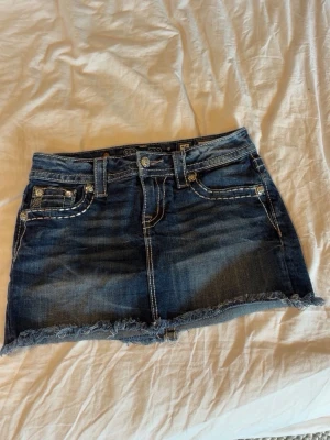 Mörkblå jeanskjol från Miss Me - Snygg mörkblå jeanskjol från Miss Me med slitna detaljer och rå kant nertill. Kjolen har dekorativa sömmar och coola broderier på bakfickorna. 