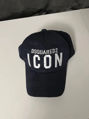 Svart keps DSQUARED2 ICON - Snygg svart keps från DSQUARED2 med stor vit brodyrtext 'ICON' framtill och märkesnamnet ovanför. Klassisk böjd skärm och justerbar baksida. Perfekt accessoar för en trendig streetwear-look.
