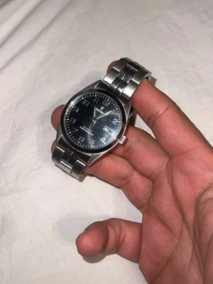 Tissot silverfärgad klocka - Stilren klocka från Tissot med silverfärgat armband i metall och svart urtavla. Klassisk rund form med tydliga siffror och datumvisning. Perfekt accessoar för dig som gillar en tidlös look.