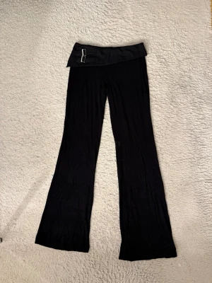 Lågmidjade yoga pants - Säljer ett par ursnygga lågmidjade yogapants med bred ribbad linning och två dekorativa spännen i midjan. Modellen har utsvängda ben och är tillverkad i ett stretchigt, mjukt material som sitter skönt på kroppen. Perfekta för en trendig och avslappnad look.