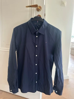 Mörkblå skjorta från Tommy Hilfiger - Snygg mörkblå skjorta från Tommy Hilfiger med klassisk krage och vita knappar. Liten broderad logga på bröstet. Skjortan har lång ärm och är tillverkad i bomull, perfekt för en stilren look.