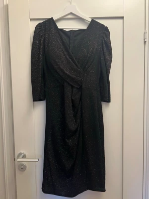 Svart glittrig omlottklänning - Svart glittrig klänning med omlott framtill och trekvartsärm. Klänningen har en elegant drapering och v-ringning, perfekt för att sticka ut. Materialet är glittrigt och ger en festlig känsla. Stängs med dragkedja bak.