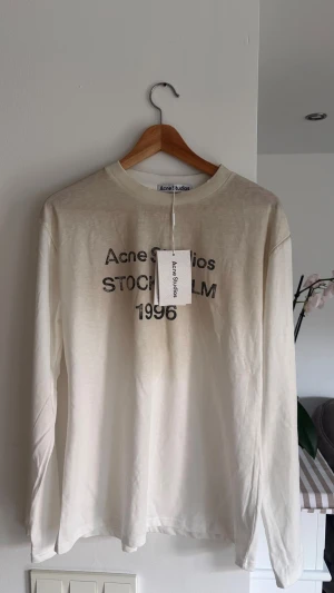 Vit långärmad topp Acne Studios - Vit långärmad topp från Acne Studios med tryck framtill där det står 'Acne Studios Stockholm 1996'. Tunn och mjuk i materialet, perfekt för en avslappnad stil. Rund halsringning och klassisk passform.