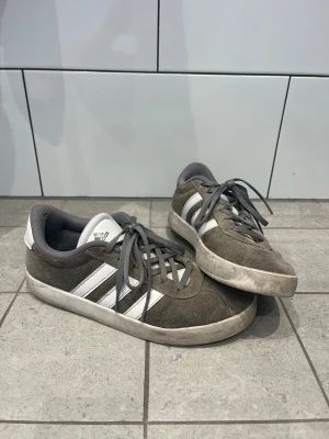 Adidas grå sneakers med vita ränder - Gråa Adidas sneakers i mocka med klassiska vita ränder på sidorna och vit sula. Skorna har lågt skaft och grå snörning. Perfekta för en avslappnad streetstil och funkar till det mesta i garderoben.