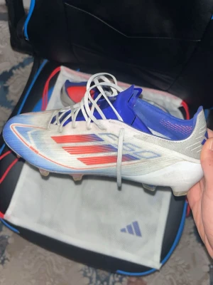 Adidas F50 fotbollsskor vit/blå/röd - Säljer ett par Adidas F50 fotbollsskor med vit bas, blå och röda detaljer samt blå innersula. Skorna har klassiska snörningar och en lätt, aerodynamisk design med plastdobbar för optimalt grepp på planen. Perfekta för dig som vill ha snabbhet och stil på matchen. Lite smutsiga bara annars så är dom perfekt inget som är förstört i skorna 