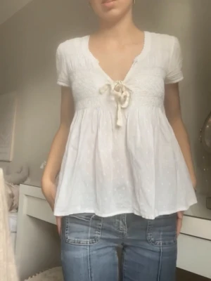 Vit blus med knyt och v-ringning - Supersöt vit blus med korta ärmar, v-ringning och knyt framtill. Blusen har en luftig passform och lätt struktur i tyget. Perfekt för en fräsch och avslappnad look.