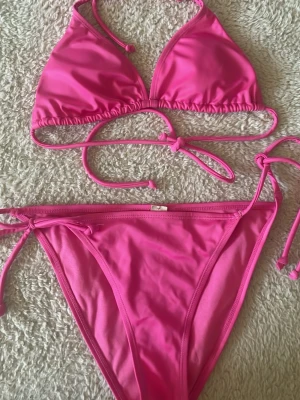 Rosa bikini med knyt i sidorna - Snygg rosa bikini med trekantsöverdel och knyt både i nacken och ryggen. Underdelen har knyt i sidorna för justerbar passform. Perfekt för stranden eller poolen. Materialet känns mjukt och följsamt.