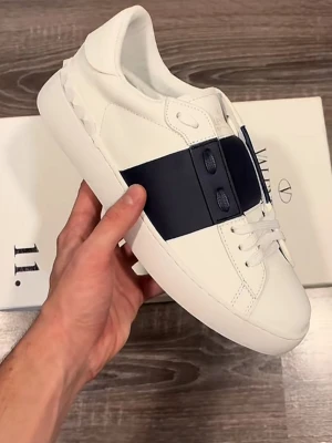 Valentino Garavani vita sneakers priset kan diskuteras  - Stilrena vita sneakers från Valentino Garavani med bred mörkblå rem över snörningen. Skorna är i läder med diskreta nitar på hälen och har en klassisk låg profil. Perfekta för dig som gillar exklusiv och modern design.