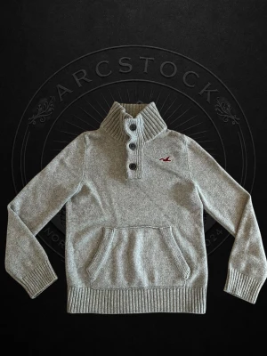 Hollister tröja - Snygg grå stickad tröja från Hollister med hög ribbad krage och tre knappar framtill. Tröjan har en magficka och Hollisters röda logga broderad på bröstet. Perfekt för kyliga dagar och enkel att matcha med jeans.
