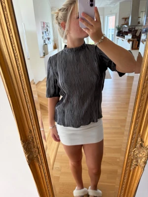 Svart plisserad blus med hög krage - Snygg svart plisserad blus med hög krage och korta ärmar. Blusen har en lös passform och ett diskret vågigt mönster i tyget. Perfekt att styla med kjol eller jeans för en trendig look.