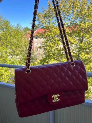 Röd quiltad axelväska  - Snygg röd axelväska  i quiltat skinn med gulddetaljer. Väskan har en klassisk design med en ikonisk logotyp på framsidan och en praktisk dragkedja inuti. Perfekt för att ge en touch av lyx till din outfit.