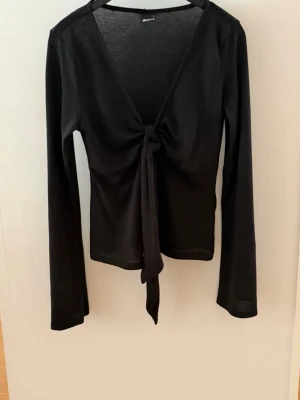 Svart långärmad knytblus Gina Tricot - Svart långärmad topp från Gina Tricot med v-ringning och knyt framtill. Tunn och stretchig modell som sitter snyggt och passar perfekt till jeans eller kjol. Enkel men trendig design som är lätt att styla.