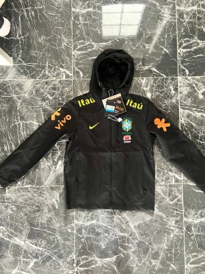 Svart Brasil jacka Nike GORE-TEX - Svart sportjacka från Nike med Brasilien CBF-logga, gula tryck på ärmar och bröst samt sponsorer som Itaú, Vivo och Guaraná Antarctica. Jackan har huva, dragkedja och är tillverkad i GORE-TEX-material som skyddar mot regn och vind.