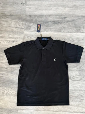 Svart pikétröja från Polo Ralph Lauren - Klassisk svart pikétröja från Polo Ralph Lauren med korta ärmar och krage. Tröjan har knäppning framtill och den ikoniska broderade loggan i vitt på bröstet. Perfekt för en stilren och avslappnad look.