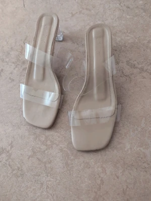 Beige sandaler med transparent klack - Stilrena sandaler i beige med fyrkantig tå och två breda transparenta remmar. Klacken är låg och genomskinlig vilket ger en modern och trendig look. Perfekta till sommarens outfits och lätta att matcha med olika stilar.
