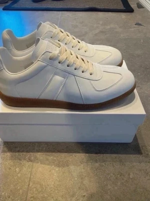 Masion Margela vita  - Snygga vita sneakers med klassisk design och beige gummisula. Skorna har ovandel i läder och mockadetaljer, samt vita skosnören. Perfekta för en clean och stilren look.44 använda en gång säljer pga försmå 