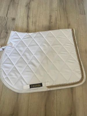 Vitt schabrak från Kerbl  - Säljer ett vitt schabrak från Kerbl PONY med quiltat mönster och dekorativt kantband i guld och svart. Schabraket har några fläckar och märken se på bild - tvättas innan de skickas eller hämtas i Sjöbo. 