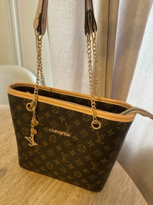  handväska - Snygg handväska  i klassiskt brunt monogrammönster med beige kanter. Väskan har guldfärgade kedjehandtag. Pris kan diskuteras 