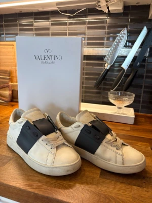 Valentino Garavani vita sneakers - Snygga vita sneakers från Valentino Garavani med bred mörk blå läderdetalj över snörningen. Klassisk låg modell med vita skosnören och diskret logga på sidan. Tillverkade i läder för en exklusiv känsla och stilren look. Priset kan diskuteras😃