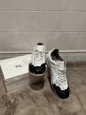 Maison Margiela vita sneakers med svart färgstänk - Maison Margiela sneakers i vitt läder med unika svarta färgstänk och svart tåparti. Klassisk låg modell med vita snören och beige sula. Snygg och edgy design som sticker ut, perfekt för dig som gillar statement-skor.  Priset kan jag pruta på, så hör av er!!