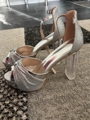 Silvriga glitterpumps med klack - Säljer ett par glittriga silvriga pumps med öppen tå, bred transparent klack och justerbar ankelrem. Skorna har metallicdetaljer och en snygg knut framtill. Perfekta för dig som vill sticka ut och glänsa på nästa event! Helt nya och oanvända. Box förekommer