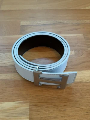 Vändbart bälte i svart och vitt med silverfärgat H-spänne - Snyggt vitt skärp med ett stort, silverfärgat H-format spänne. Perfekt accessoar för att lyfta din outfit med en clean och modern look, funkar till allt. Bältet är 110cm och i mycket bra skick✅ Fraktar inom en dag✅ Priset är inte hugget i sten🙂