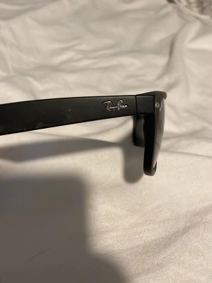 Svarta Ray-Ban solglasögon  - Klassiska svarta Wayfarer solglasögon från Ray-Ban med mörka glas och ikonisk logga på bågen. Snygg och tidlös design som passar till allt. Bågarna är i matt svart plast och har en rak, stilren form.
