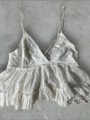 Vit volangtop med spetsdetaljer - Söt vit topp med tunna axelband, volanger och spetsdetaljer framtill. Toppen har v-ringning och smock i ryggen för skön passform. Materialet är tunt och lätt, perfekt för sommardagar. Romantisk och bohemisk stil som passar till jeans eller kjol.