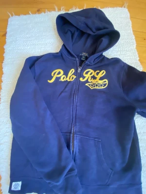Marinblå hoodie från Polo Ralph Lauren - Snygg marinblå hoodie från Polo Ralph Lauren med gul broderad text 'Polo RL' och stjärndetalj på bröstet. Dragkedja framtill, två fickor och klassisk huva. Perfekt för en avslappnad och sportig stil.
