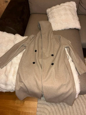 Beige ullkappa med huva - Stilren beige kappa i ullmix med stor huva och dubbelknäppning framtill med svarta knappar. Lång modell med raka ärmar och diskreta fickor på sidorna. Perfekt för dig som vill ha en klassisk men ändå modern look.