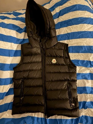 Svart dunväst med huva från Moncler - Snygg svart dunväst från Moncler med avtagbar huva och två dragkedjefickor framtill. Säljer pga för liten, använd 1 gång!  Dragkedjan på högra fickan är sönder annars är allt bra. Pris går att diskutera