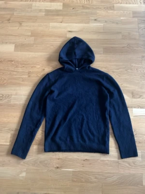 Mörkblå hoodie i mjukt material - En stilren mörkblå hoodie med huva och långa ärmar. Tröjan är tillverkad i ett mjukt, stickat material som känns skönt mot huden. Perfekt för dig som gillar en enkel och clean look. Ribbstickade muddar vid ärmslut och nederkant ger en snygg passform.